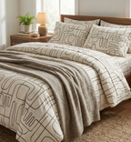 The image displays a cozy bedroom scene featuring a large bed with a distinctive bedding set. The duvet cover and pillowcases have an abstract, continuous black line pattern on a light background. A textured, light brown or taupe throw blanket is draped across the foot of the bed.