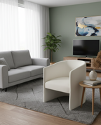 A light grey two-seater sofa with a throw pillow is on the left. In the foreground on the right is a white, curved armchair. A light wood coffee table sits next to the armchair, holding a white vase and a stack of books.