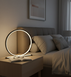 Modern circular LED lamp, open book, and glasses on a wooden nightstand in a dark bedroom.