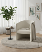 a modern, minimalist living space or corner featuring a cream-colored armchair as the focal point. The chair, possibly an Elmore Chair by Four Hands, features a unique "shapely, fold-style form" with a barrel back and sculpted open base. A light-colored throw blanket is draped over one arm. 